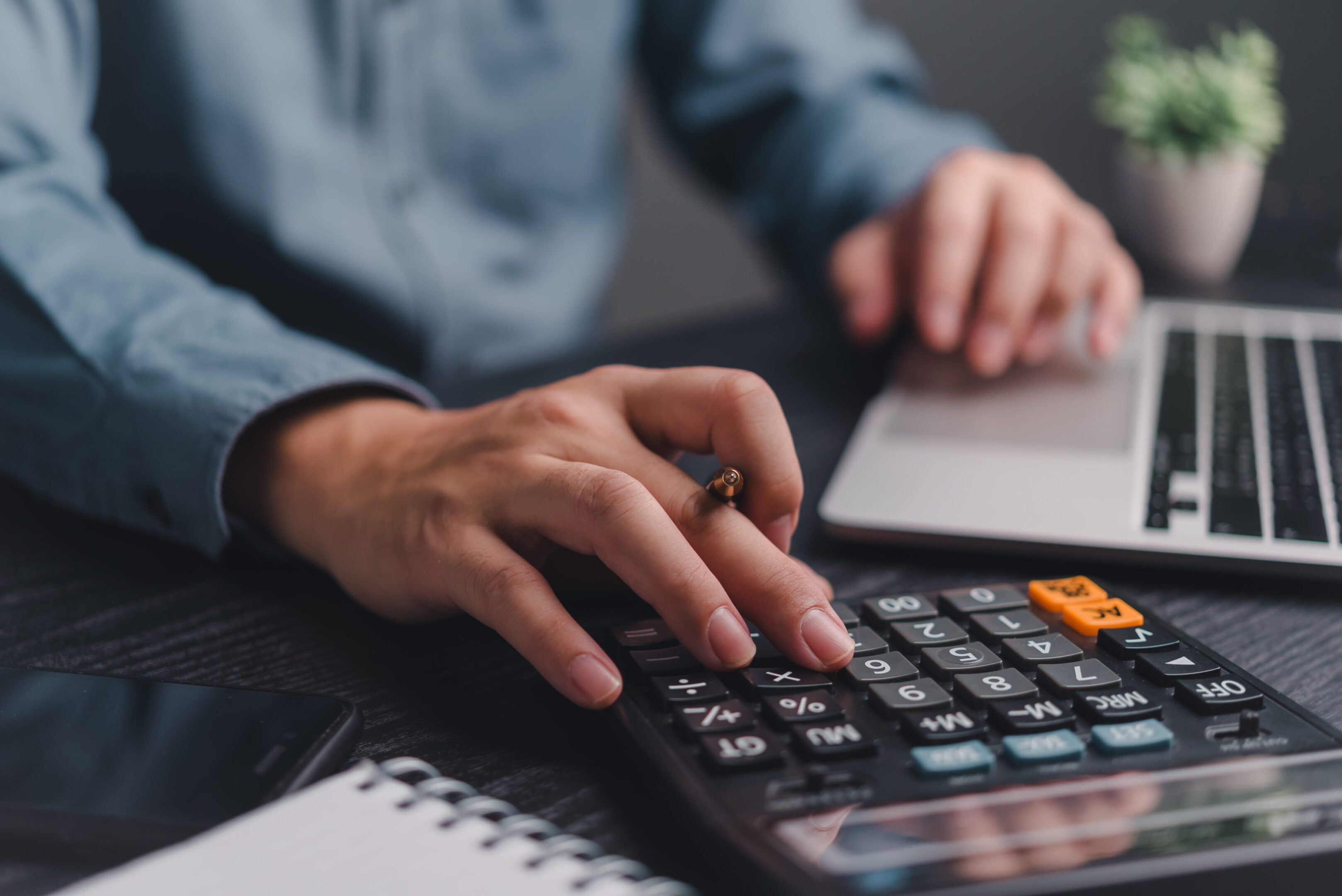 Person using calculator and laptop at desk.